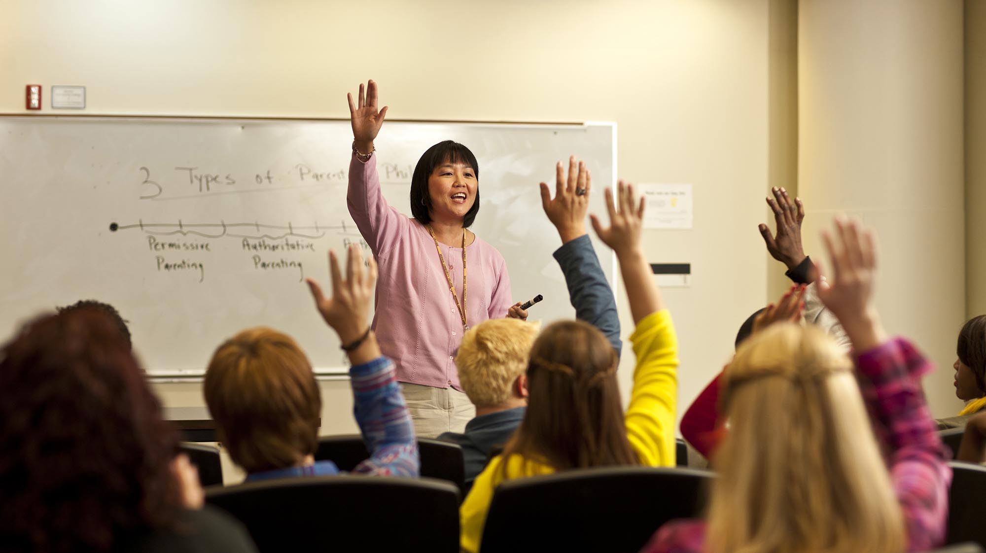 A teacher stands at the front of a classroom while several students raise their hands to answer a question. A whiteboard with handwritten notes is visible behind the teacher.