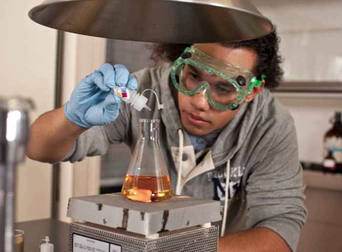 An MCC student wearing goggles conducts an experiment