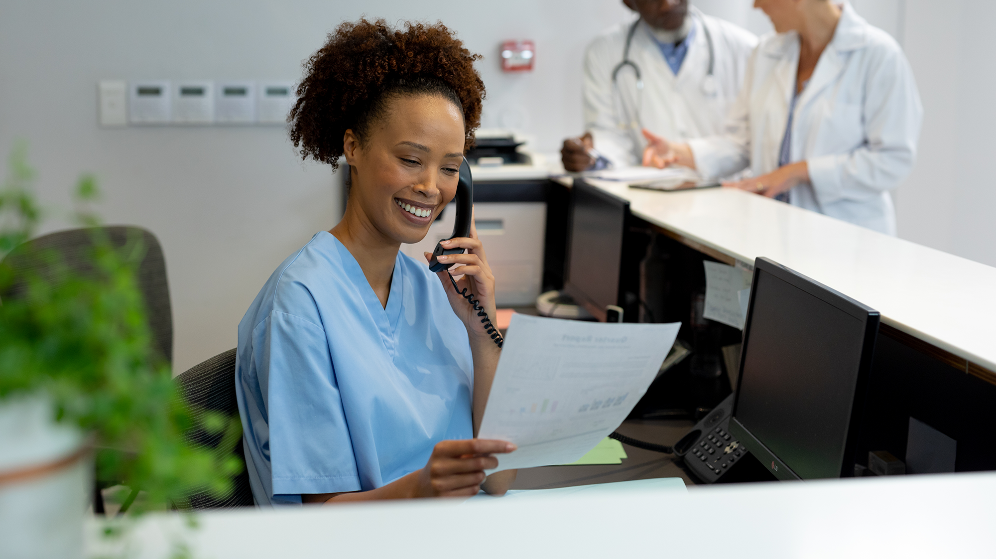 a woman wearing scrubs sitting at a desk answering a phone.