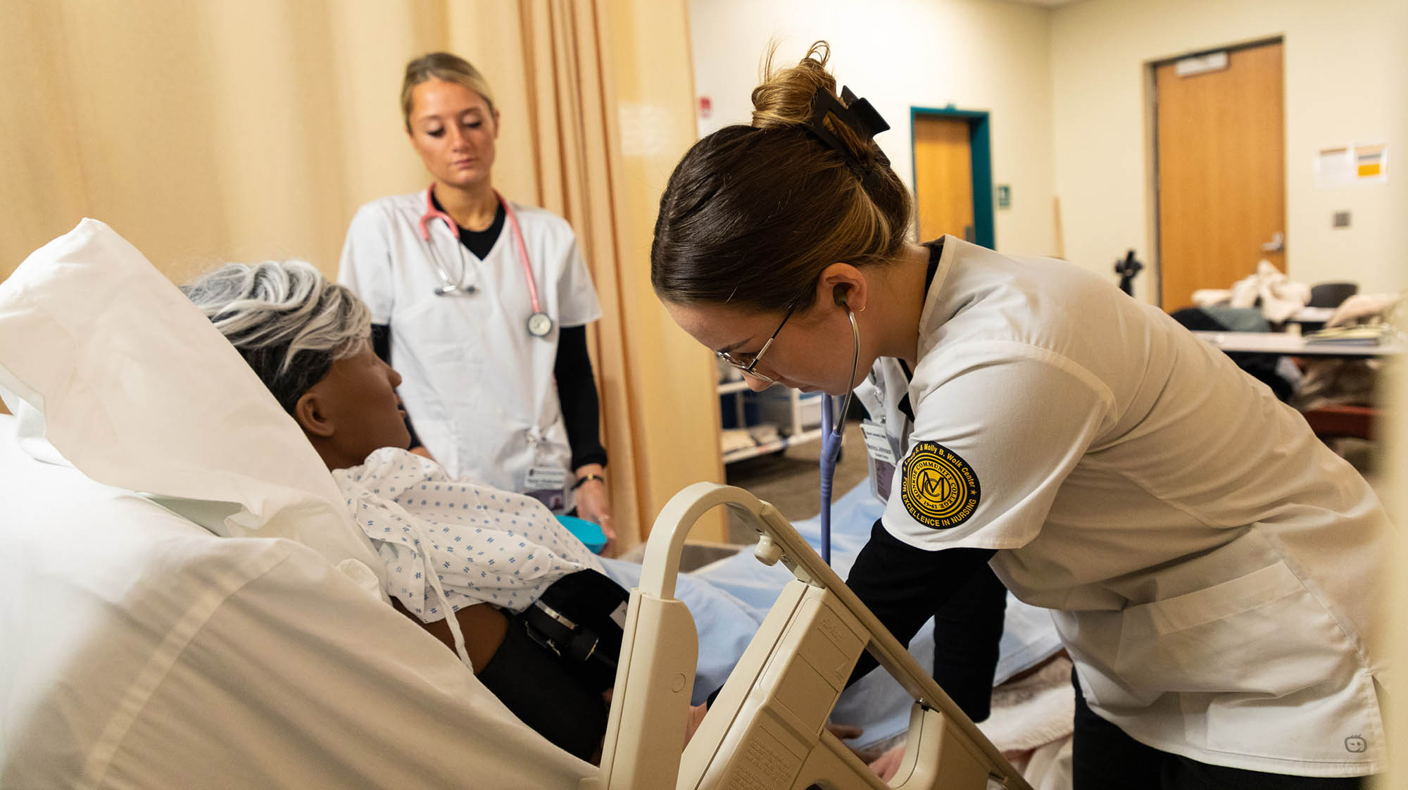 Nursing students provide hands-on patient care in a simulated hospital setting, with one student examining a patient mannequin while another observes during a clinical skills training session.