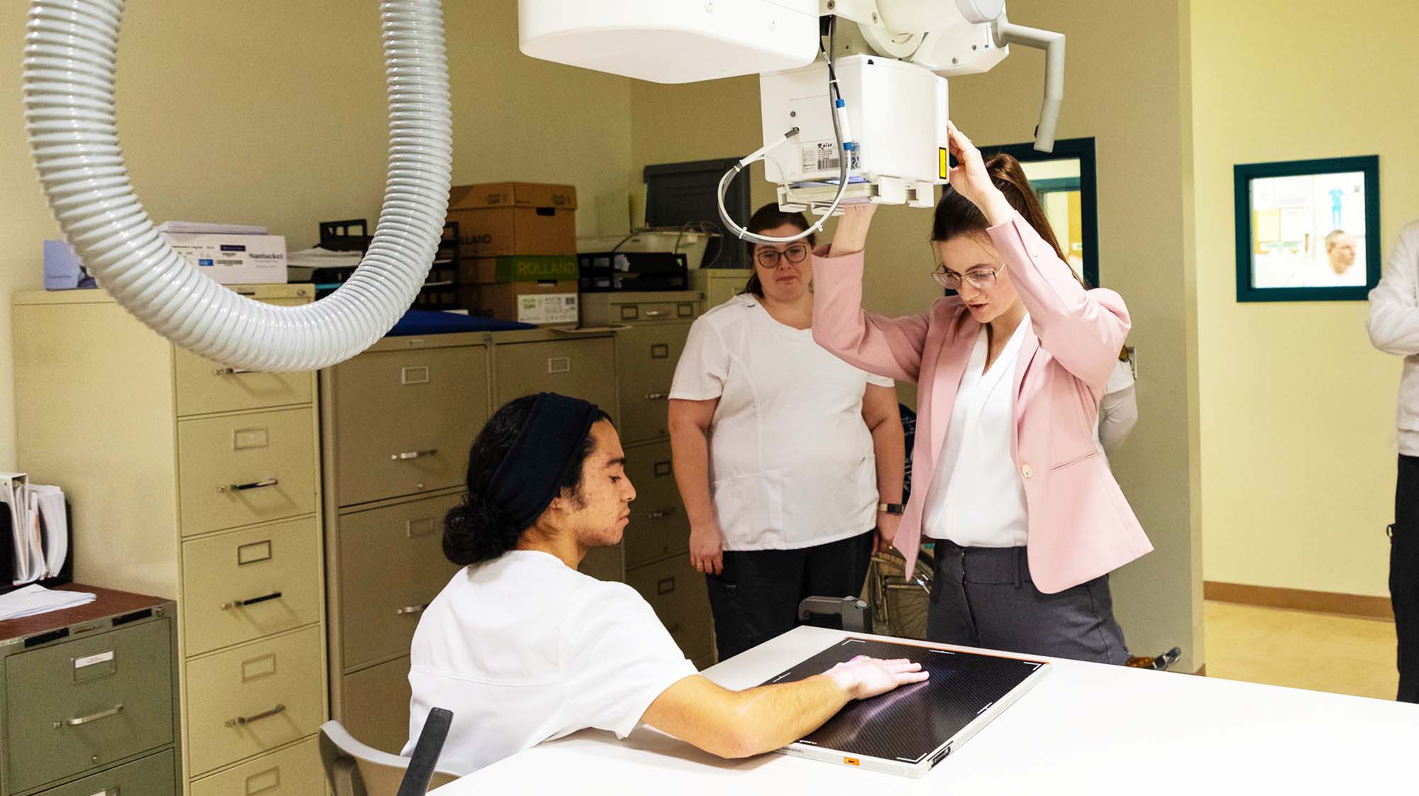 Radiologic technology student positions an X-ray machine while another student serves as the patient during a hands-on lab exercise.