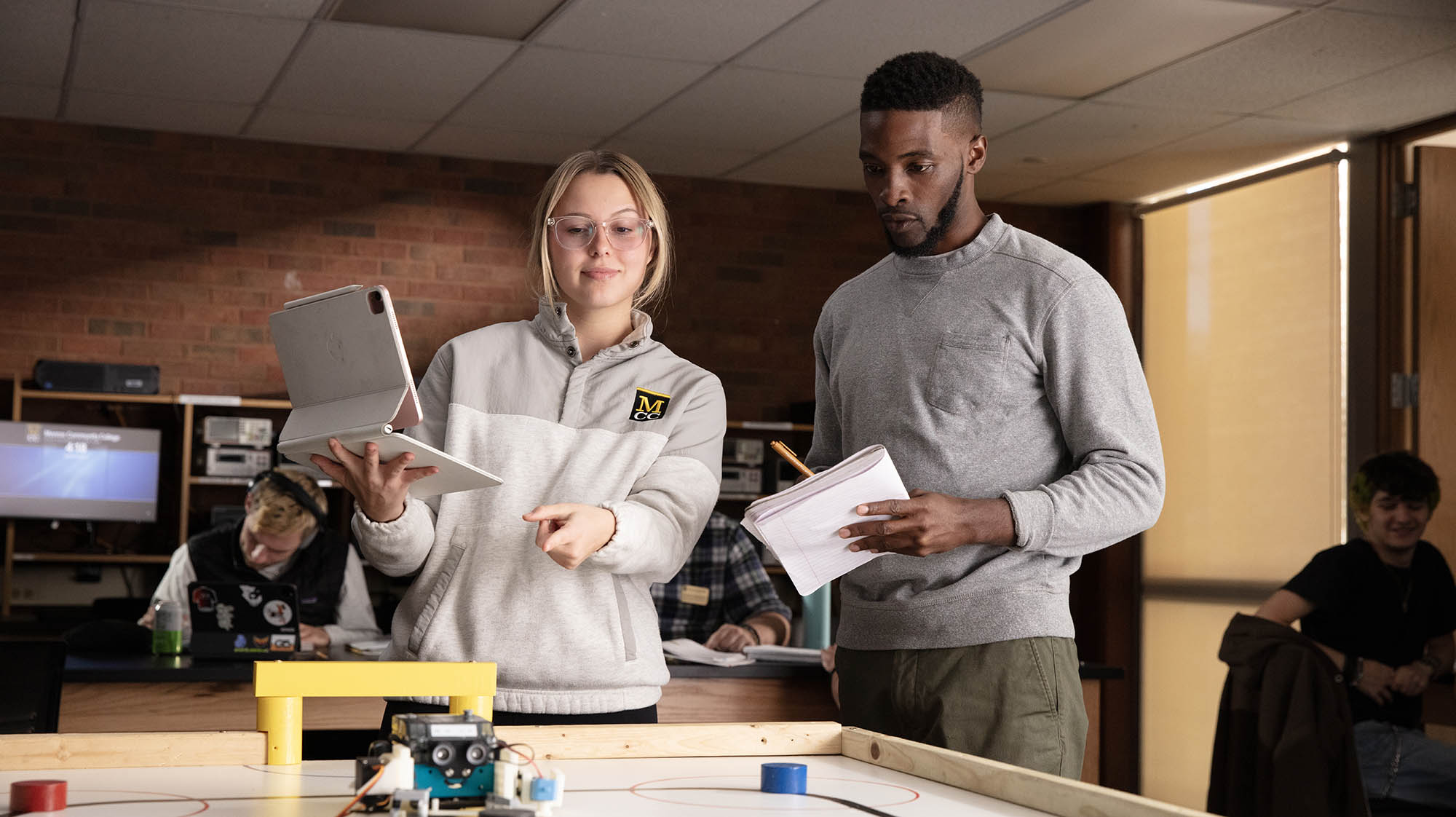 Two MCC students collaborate on a robotics project in a classroom, with one student holding a tablet and pointing toward a tabletop robot while another takes notes.