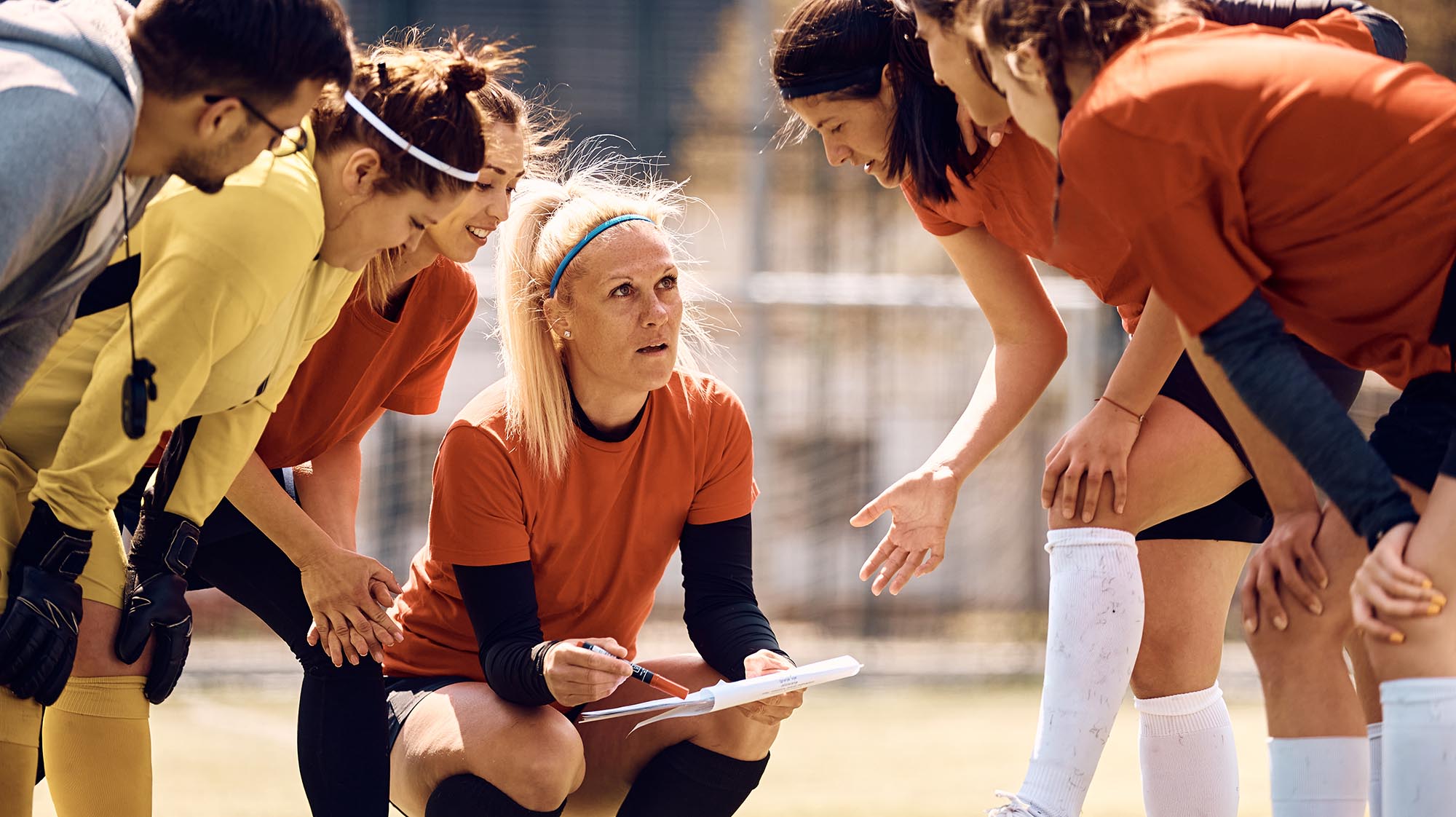 Coach kneels with a clipboard while leading a strategy discussion with student athletes during an outdoor team practice.