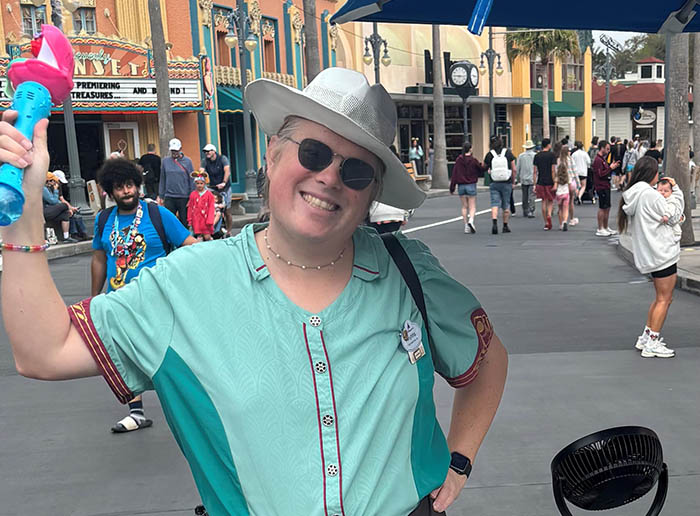 a person working in disney world wearing a teal shirt and a white fedora