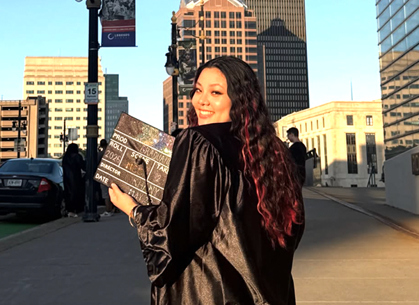 a recent graduate posing with her cap and gown, a cityscape is seen in the background
