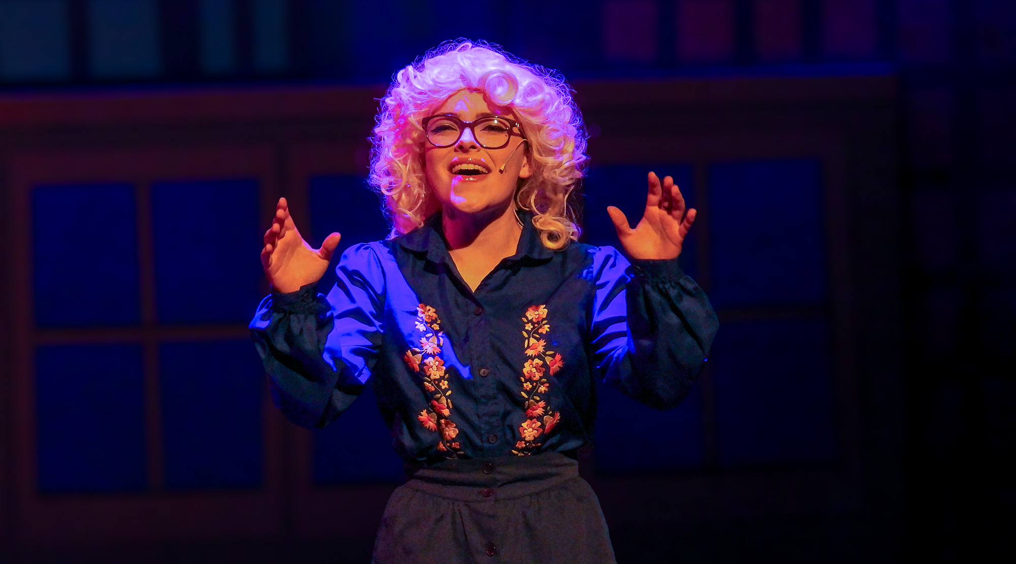 A performer wearing a curly white wig, glasses, and a dark embroidered blouse stands on stage under vivid blue and purple lighting, smiling and gesturing with both hands. A dimly lit theater backdrop with dark blue panels fills the background.