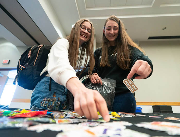 Two students smiling and reaching for items on a table at a graduation fair.