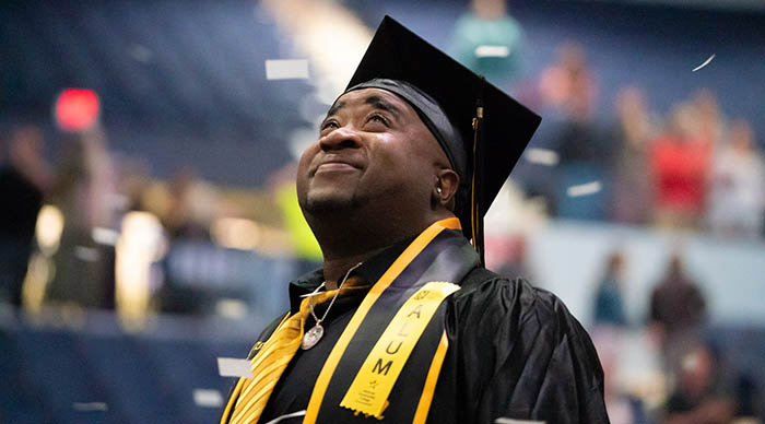 A smiling Monroe Community College graduate in cap and gown looks upward as confetti falls during commencement.