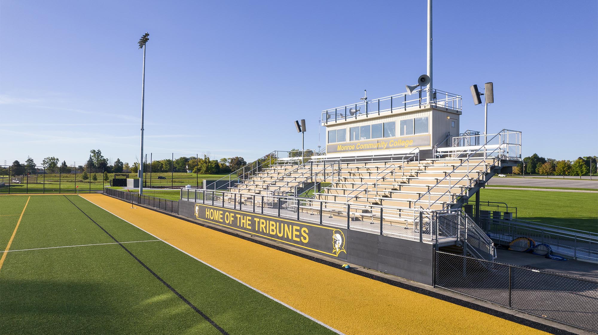 Exterior view of Monroe Community College’s athletic field and bleachers with “Home of the Tribunes” signage.